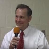 White man with brown hair, white shirt and red tie talking into a microphone