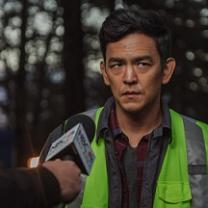 Man with dark hair and a yellow vest with a news media microphone aimed at him and he has a worried look on his face