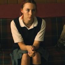 Young white woman in a white blouse, vest and hair pulled back sitting on a couch with hands clasped looking irritated