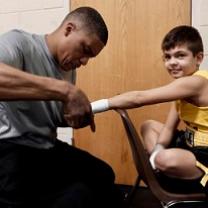 Black man in gray shirt taping up the hand of a young white boy in a yellow tank top