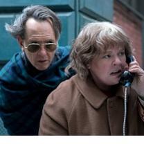 A man with sunglasses and hair blowing in the wind hovering behind a woman looking upset talking into a pay phone