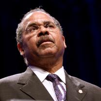 Black man in a suit with glasses speaking with his head tilted back