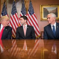 Trump, Ryan and Pence at a table looking at each other with flags in the back