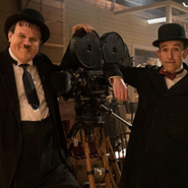 One large and one small man both in black suits with white shirts and hats leaning on an old time film projector