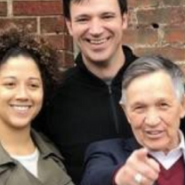 Young black woman smiling on the left, tall white man smiling in the middle and older white man pointing at the camera standing in front of a brick wall