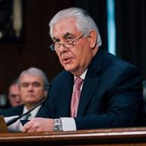Older man with gray hair, a dark suit and pink tie sitting at a mic with glasses halfway down his nose