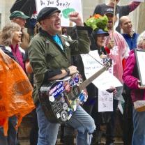 Guy with cap and guitar covered with stickers with his fist in the air in front of a crowd of people