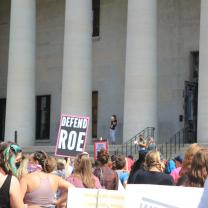 crowd protesting in front of state house with sign that says defend roe